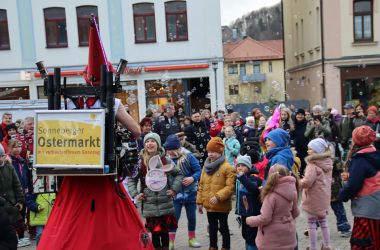 Ein rot kostümierter Mann trägt ein Schild auf dem Rücken mit der Aufschrift: Sonneberger Ostermarkt mit verkaufsoffenem Sonntag. Mit einem Gerät erzeugt er Seifenblasen. Um ihn herum versammeln sich viele Kinder.
